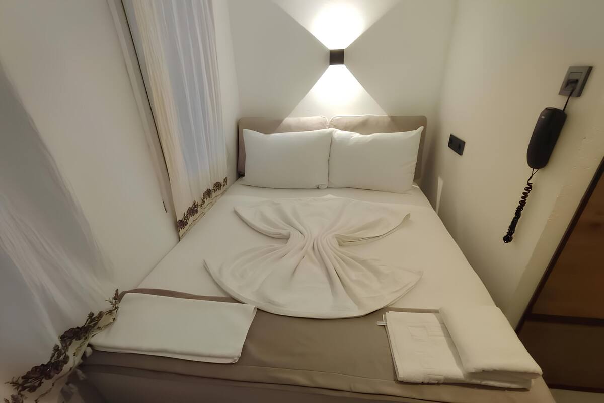 A neatly made bed with a folded white towel and a neatly folded white blanket. There is a wall-mounted phone and a light fixture above the bed.