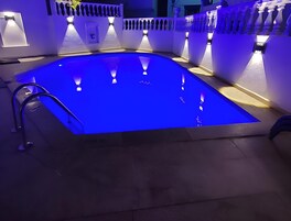 Una piscina con un tono azul, rodeada de paredes y columnas blancas, e iluminada por luces colgantes.