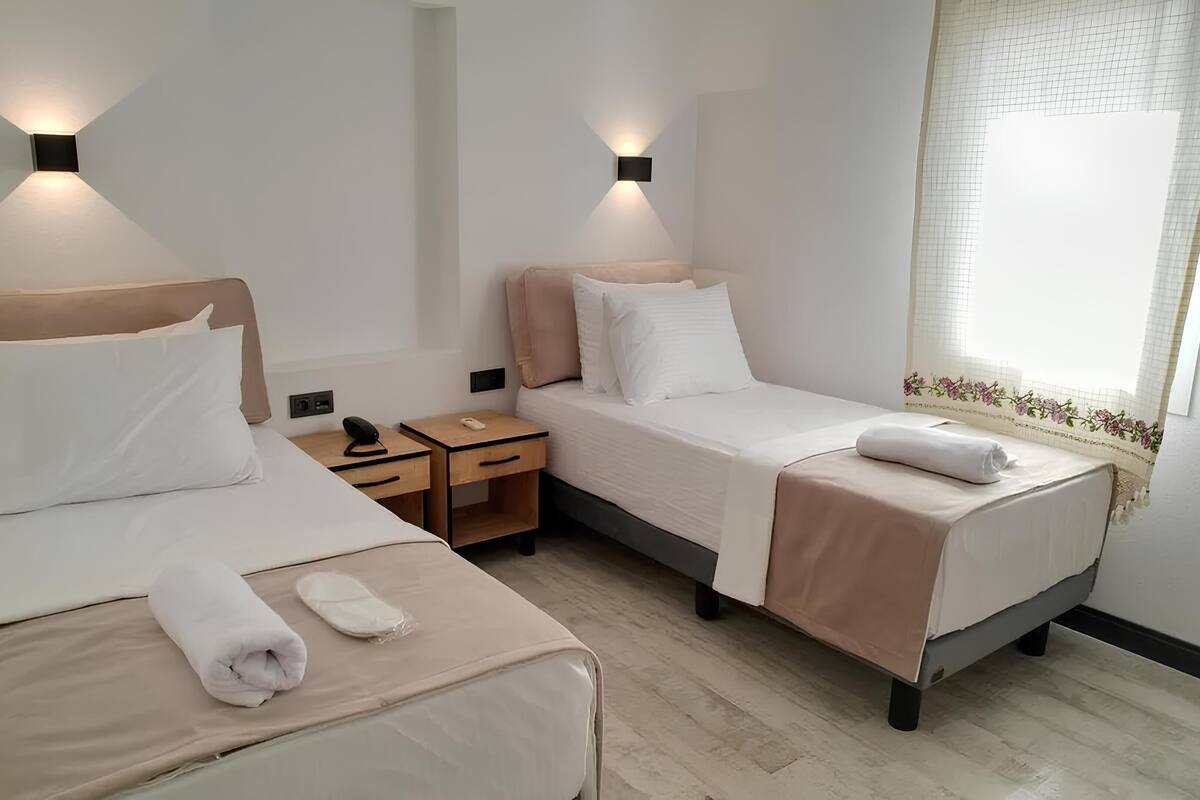 A hotel room with two single beds, bedside tables, wall-mounted lamps, a window with a floral curtain, and a wooden floor.