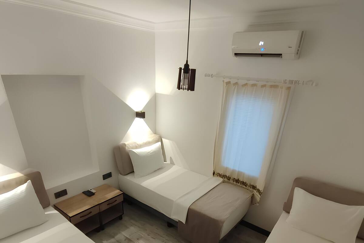 A hotel room with three single beds, a bedside table with a lamp, a window with curtains, and an air conditioning unit.