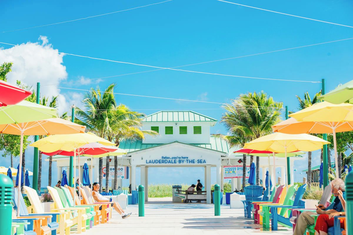 A beachside pavilion with colorful umbrellas and lounge chairs, a sign reading 'LAUDERDALE BY THE SEA', and a clear view of the sea.