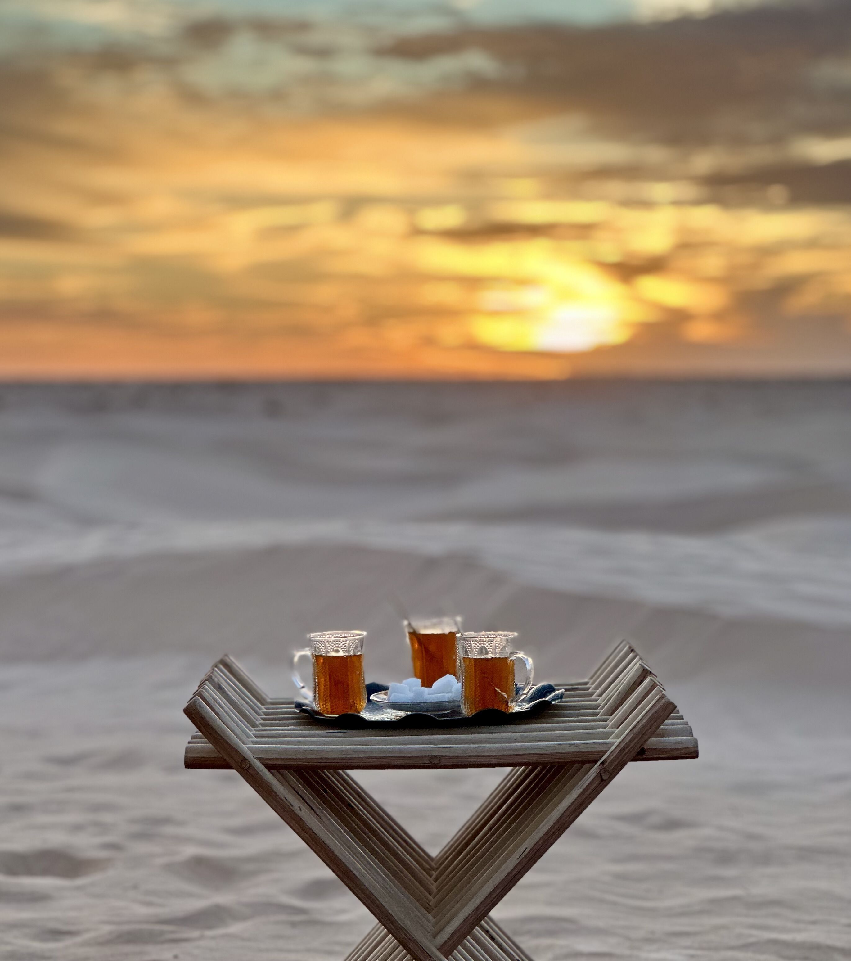 Ein Holztisch mit drei Teetassen und Zuckerwürfeln an einem Strand bei Sonnenuntergang.