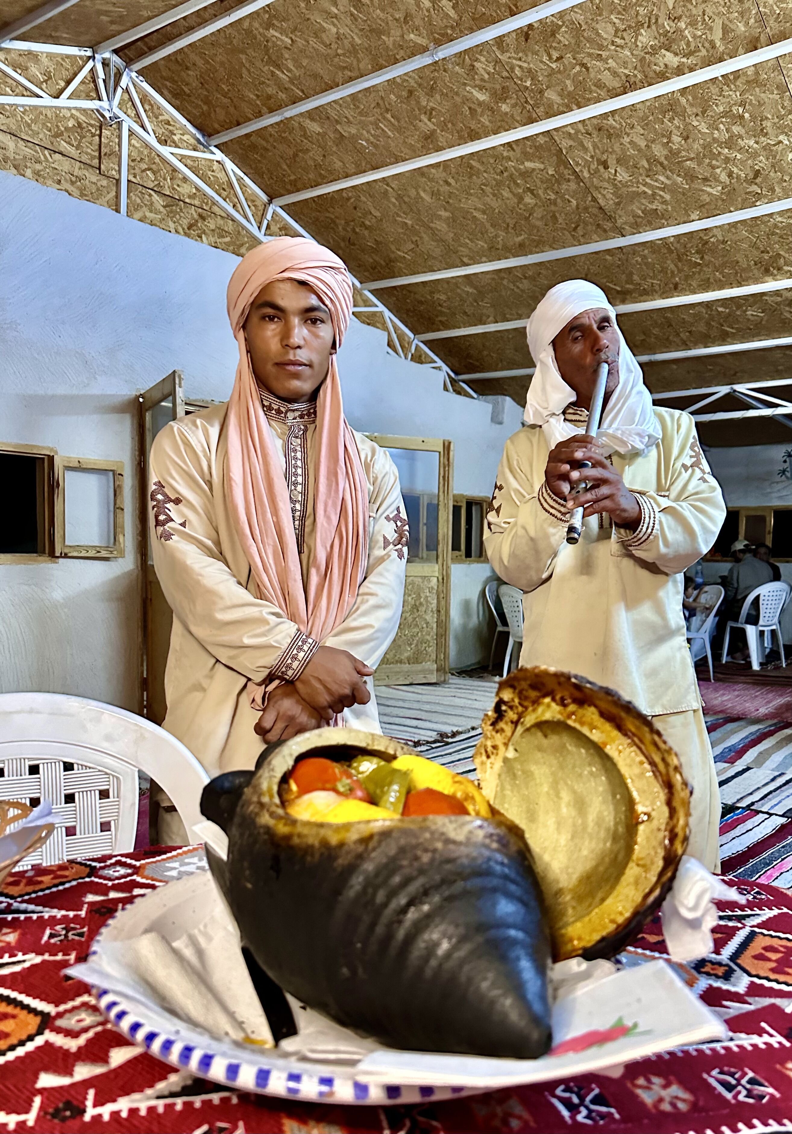 Two individuals in traditional attire standing in a room with a patterned carpet, a large bowl with food, and a white chair in the background.