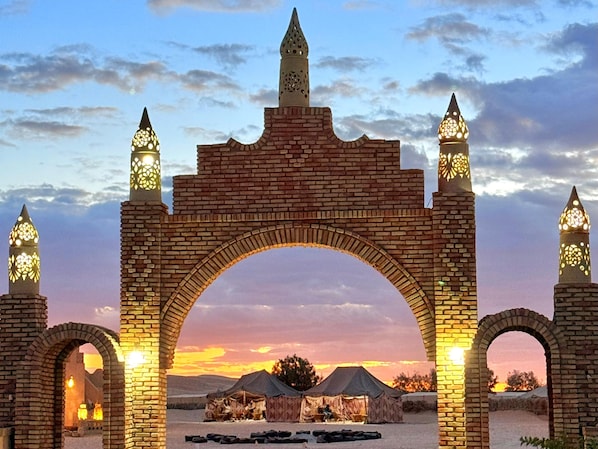 A brick archway with four lanterns, leading to a campsite with tents and a clear sky.