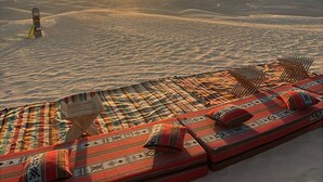 Woestijnlandschap met een ondergaande zon, een loungeruimte met gestreepte kussens, en een snowboard op de achtergrond.