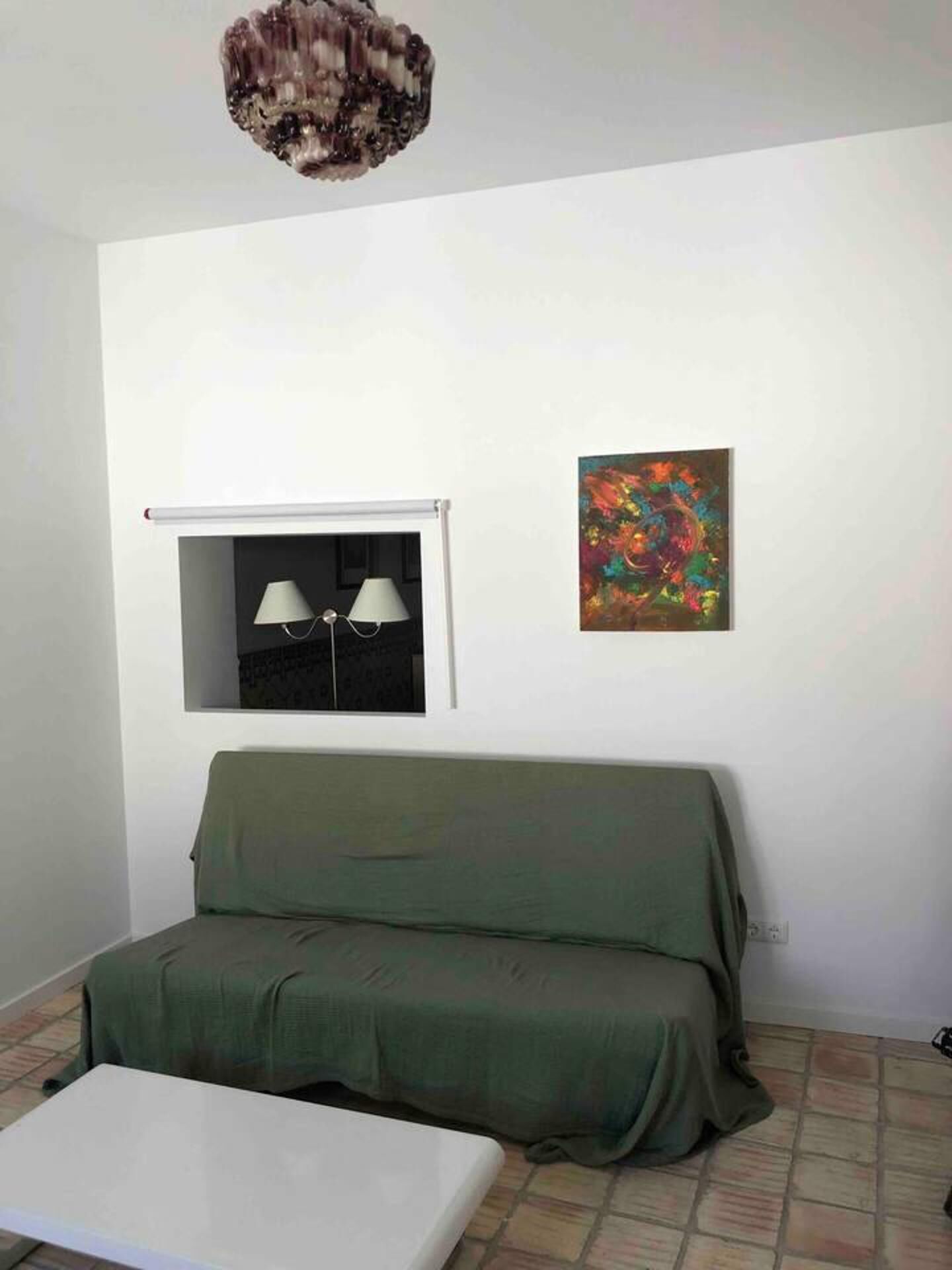 Una habitación con un sofá verde, una mesa blanca, un ventanal con dos lámparas y un cuadro abstracto colorido en la pared.