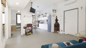 Een moderne woonkamer met een bank, een eethoek met stoelen, een keuken met een koelkast en een televisie die aan de muur is bevestigd.