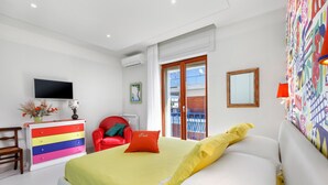 A bedroom with a bed, a colorful dresser, a red chair, a television, and a balcony view. - Estate4home - Esé (Sorrento)