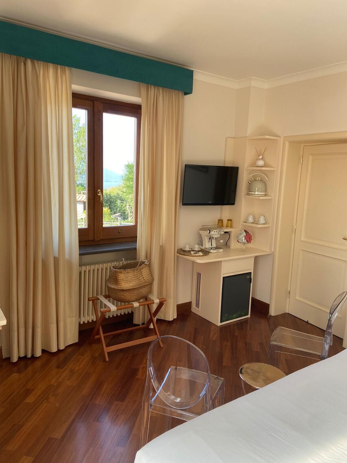 Habitación de hotel con piso de madera, una cama, un escritorio con televisor, un radiador, una ventana con cortinas y vista a zonas verdes.