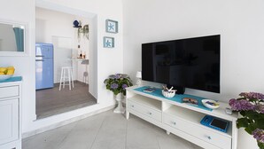 Un salon moderne doté d’un téléviseur à écran plat, d’un meuble de rangement blanc et d’un coin cuisine visible à travers une porte ouverte.