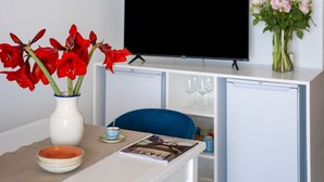 A room with a white table, a white cabinet with a television, a vase with red flowers, and a vase with white flowers. - Estate4home - Emeri Positano (Positano)
