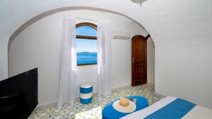 A room with a TV, a window with a view of the sea, a wooden door, and a ceiling light. - Estate4home - Emeri Positano (Positano)