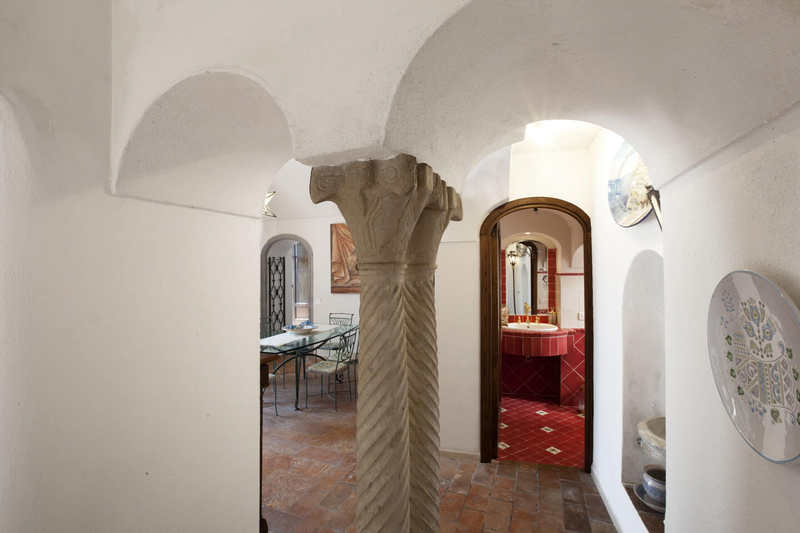 A hallway with a stone column, a dining area with a table and chairs, and a red-tiled bathroom.