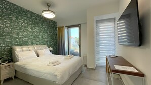 A neatly made bed with white linens and a folded towel, a bedside table with a lamp, a wall-mounted TV, and a window with blinds. - AirTLV - Luxury Apartment Port of Jaffa (Tel Aviv)