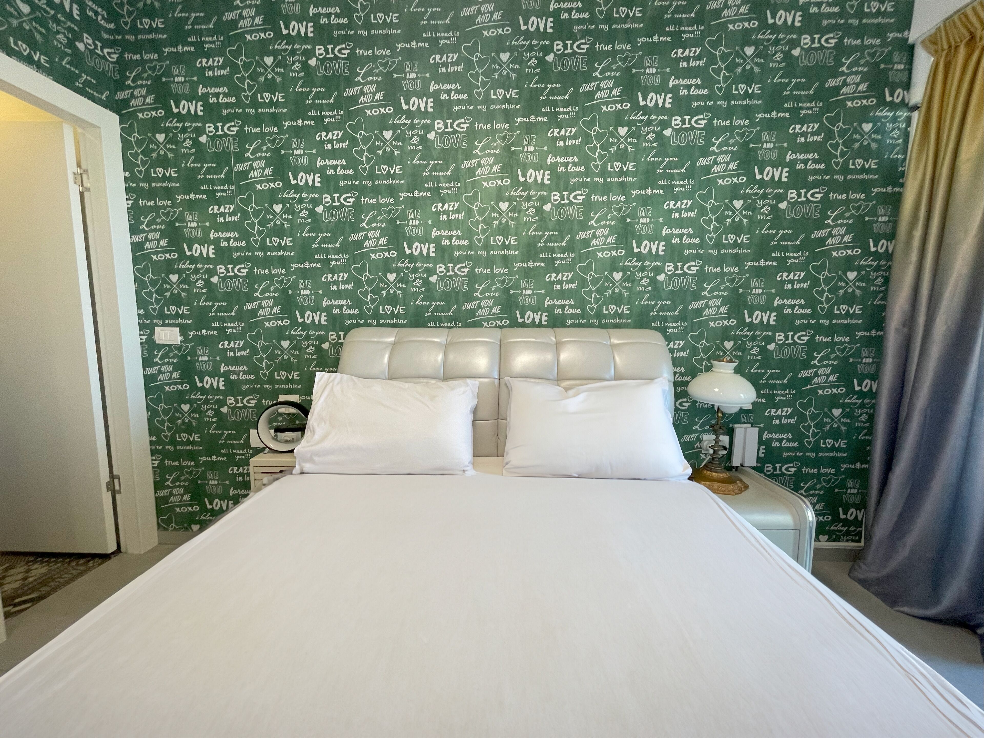 Un dormitorio con una pared verde cubierta por las palabras 'LOVE' y 'BIG', una cama blanca con dos almohadas y una mesita de noche con una lámpara.