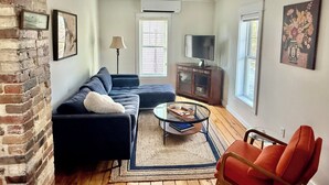 A living room with a sectional sofa, a glass coffee table, a red armchair, a TV on a cabinet, and a brick wall. - Cozy 1+ Bedroom Haven with Charm and is a block from downtown! (Manistee)