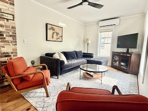 A living room with a sectional sofa, a red armchair, a glass coffee table, a brick wall, a ceiling fan, and a TV on a wooden cabinet.