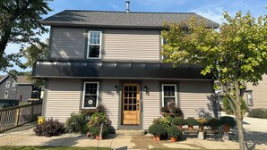 A two-story house with a front door, windows, and a small porch area with potted plants. - Cozy 1+ Bedroom Haven with Charm and is a block from downtown! (Manistee)