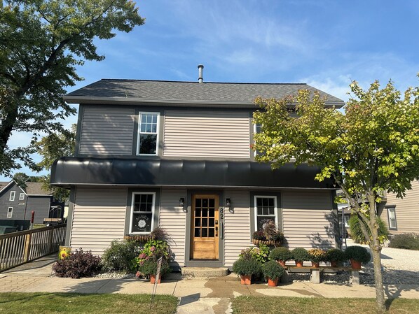 A two-story house with a front door, windows, and a small porch area with potted plants.