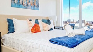 A hotel room with a large bed, multiple pillows, a view of the city through large windows, and a painting on the wall. - Union Towers Condo with City Views and Patio (Kitchener)