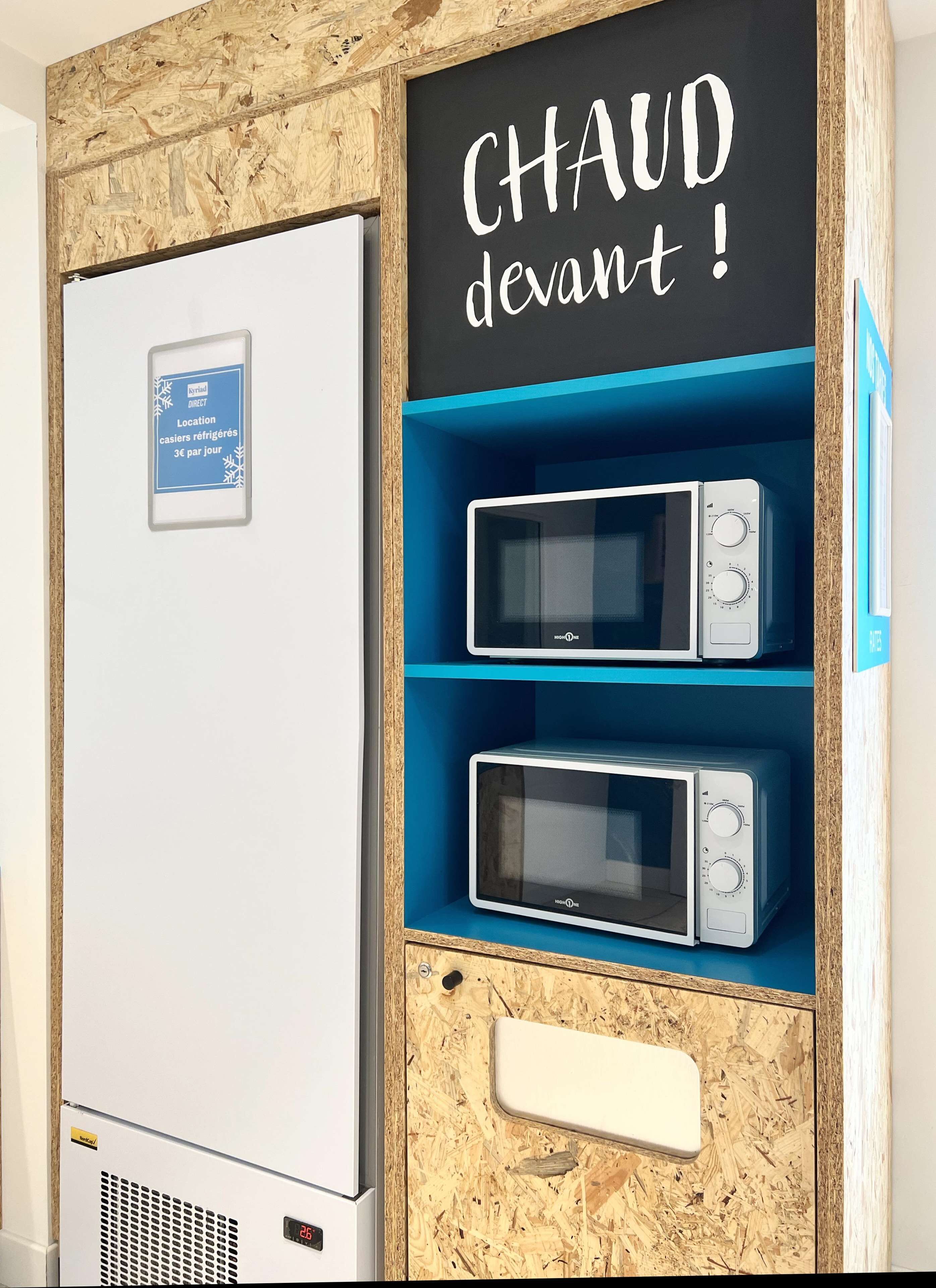 A kitchenette with a white refrigerator, two microwave ovens on blue shelves, and a wooden cabinet with a chalkboard sign saying "CHAUD devant!".