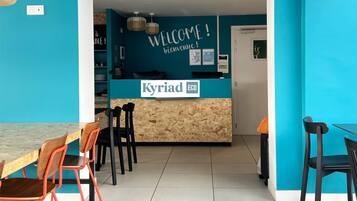 Un área de recepción con pared azul, un mostrador de madera y un letrero que dice "Kyriad ECO". Se ven sillas y una mesa en primer plano.