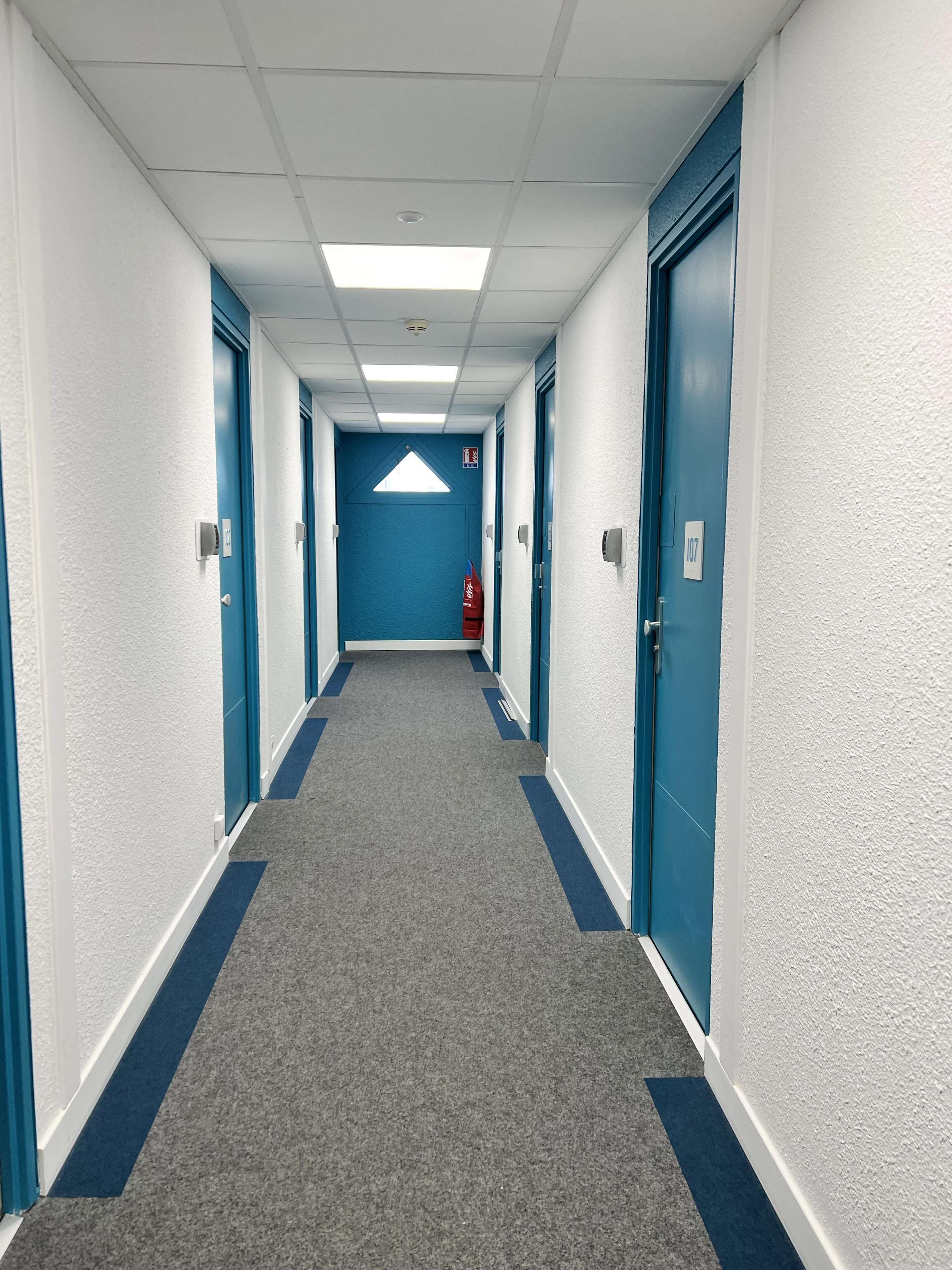 A long, narrow hallway with blue doors and white walls, leading to a room marked with the number 107.