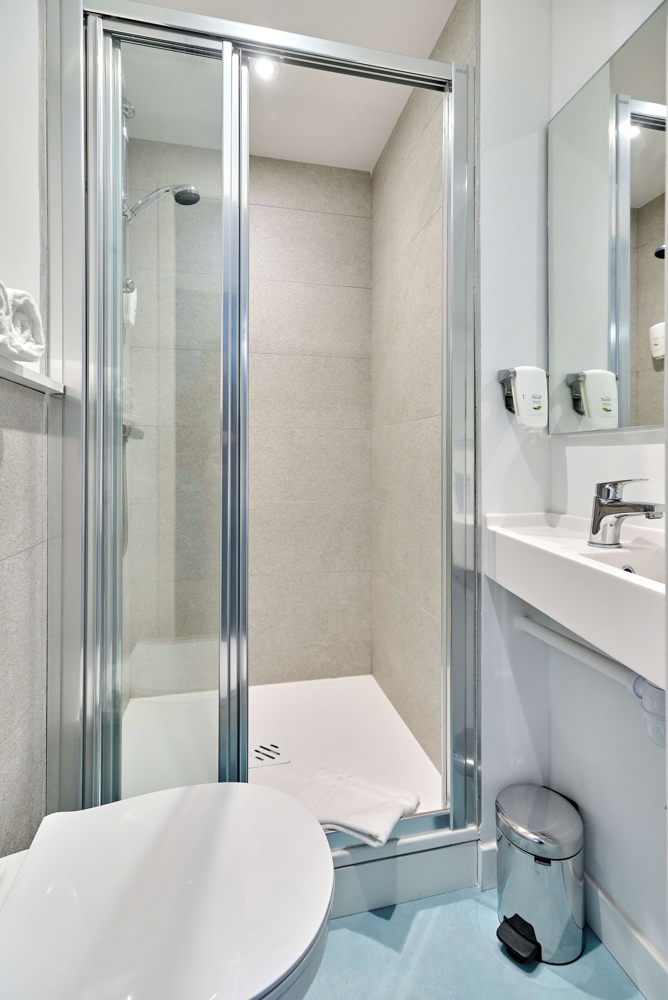 A modern bathroom with a glass-enclosed shower, a white sink, a mirror, and a trash bin.