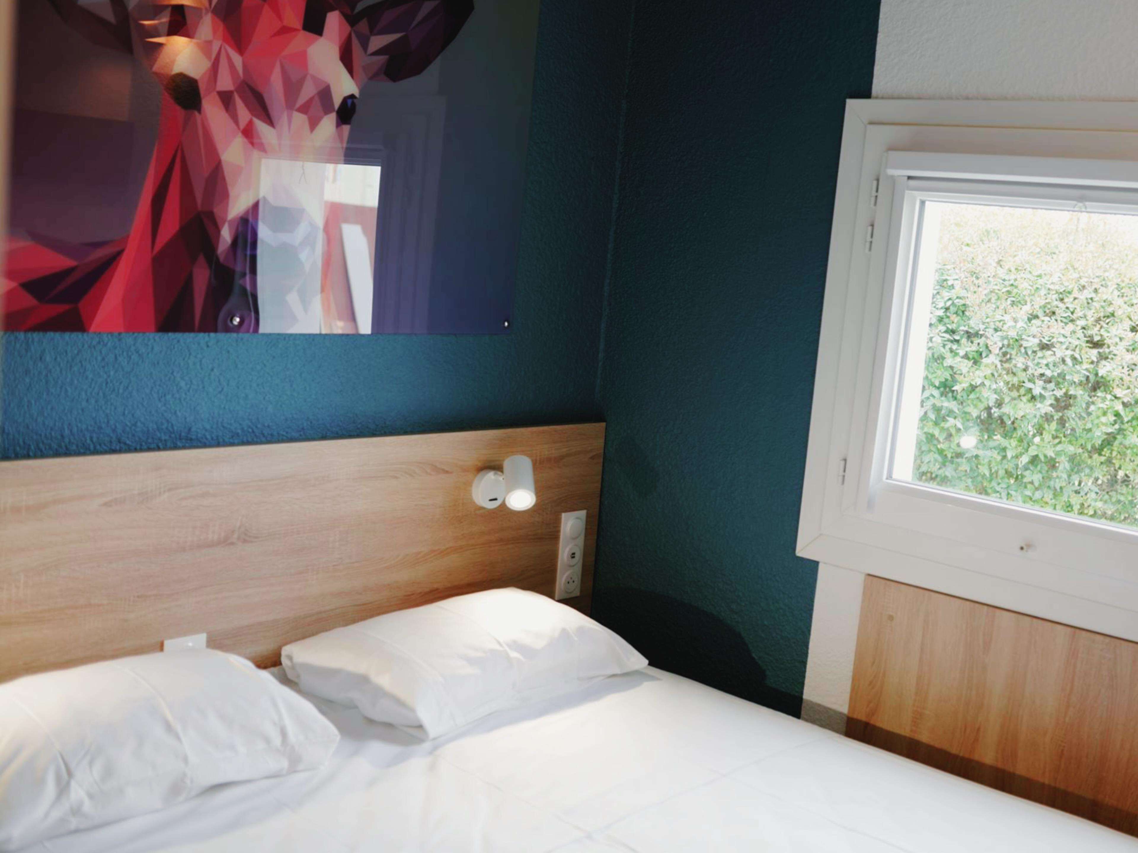 Habitación de hotel con una cama, cabecero de madera, una lámpara de pared y una ventana con vistas al verdor.