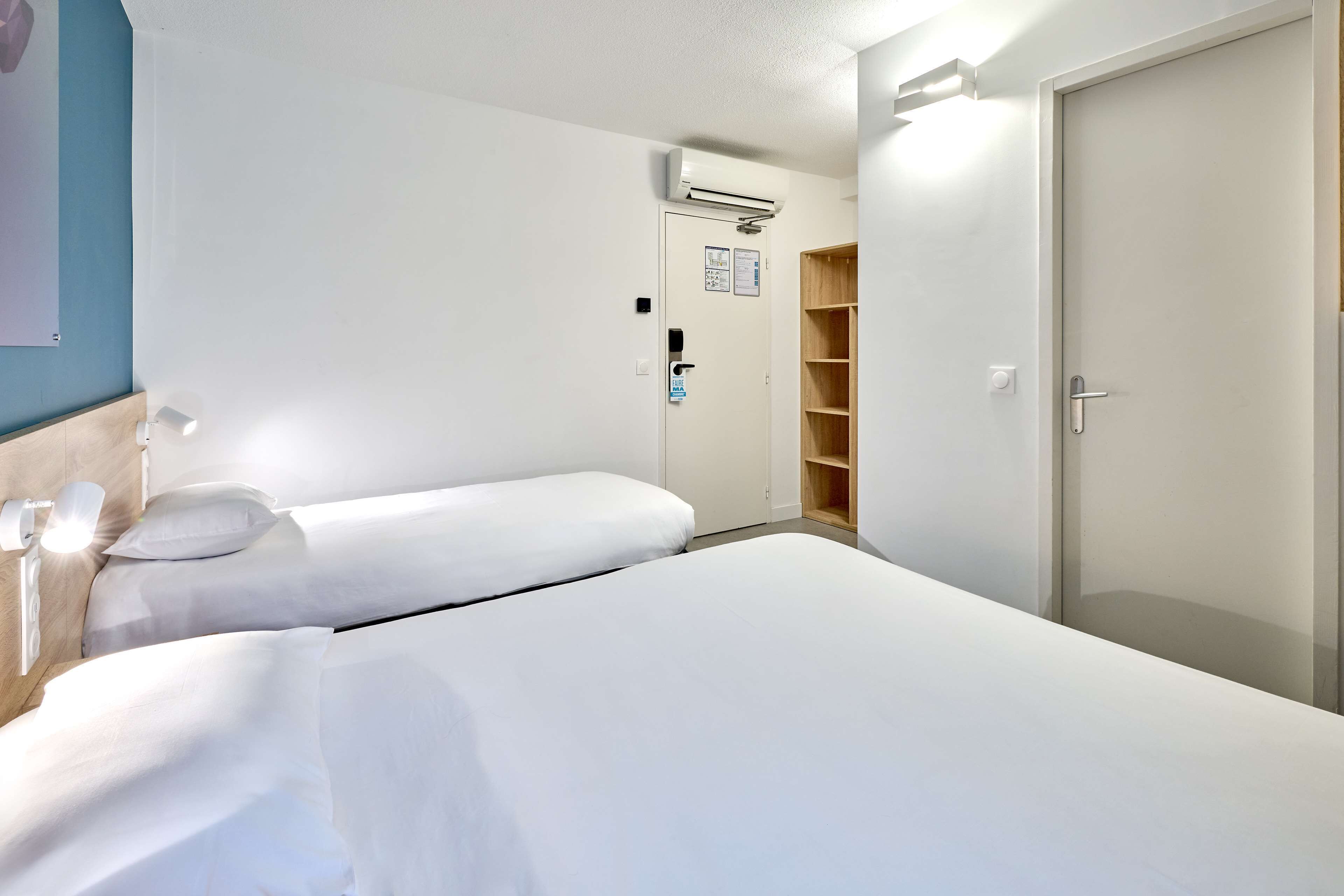A hotel room with two single beds, a wardrobe, an air conditioning unit, and a door with a handle.