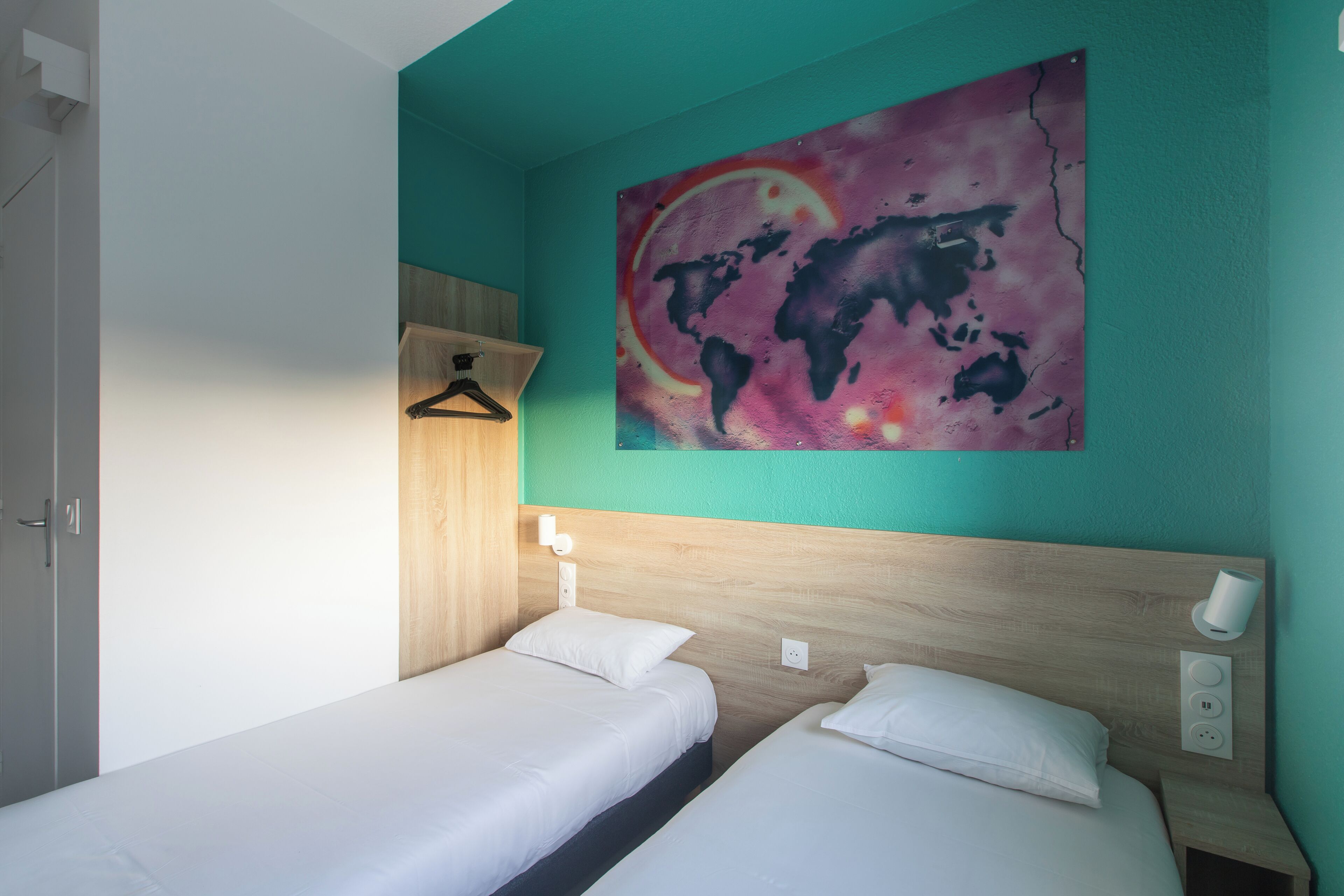 A room with two beds, a wooden headboard, a world map painting, a light switch, and a hanging light fixture.