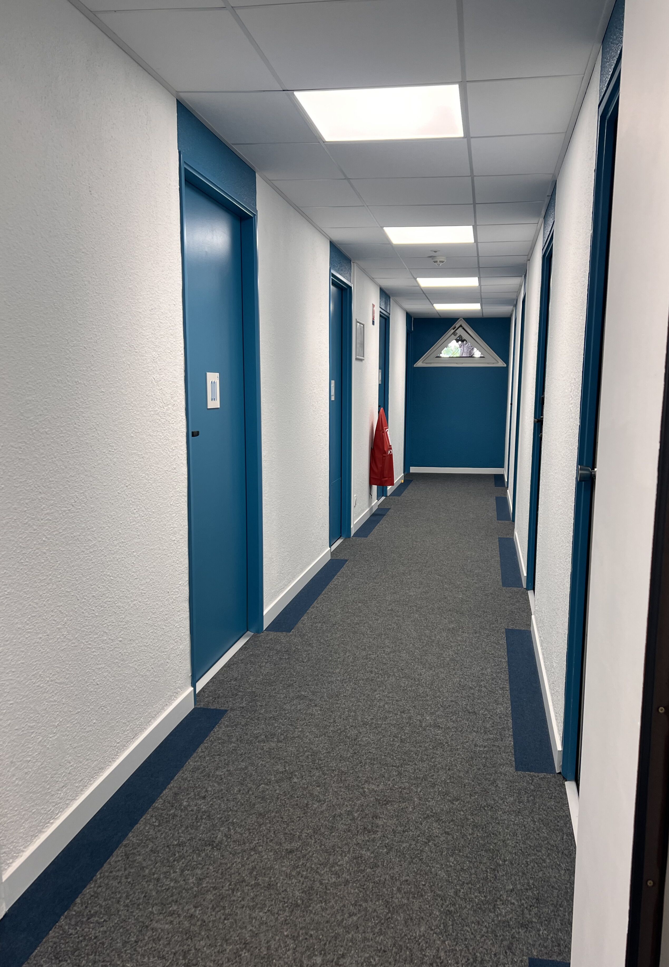 A long, narrow hallway with blue doors and white walls, carpeted floor, and overhead fluorescent lighting.