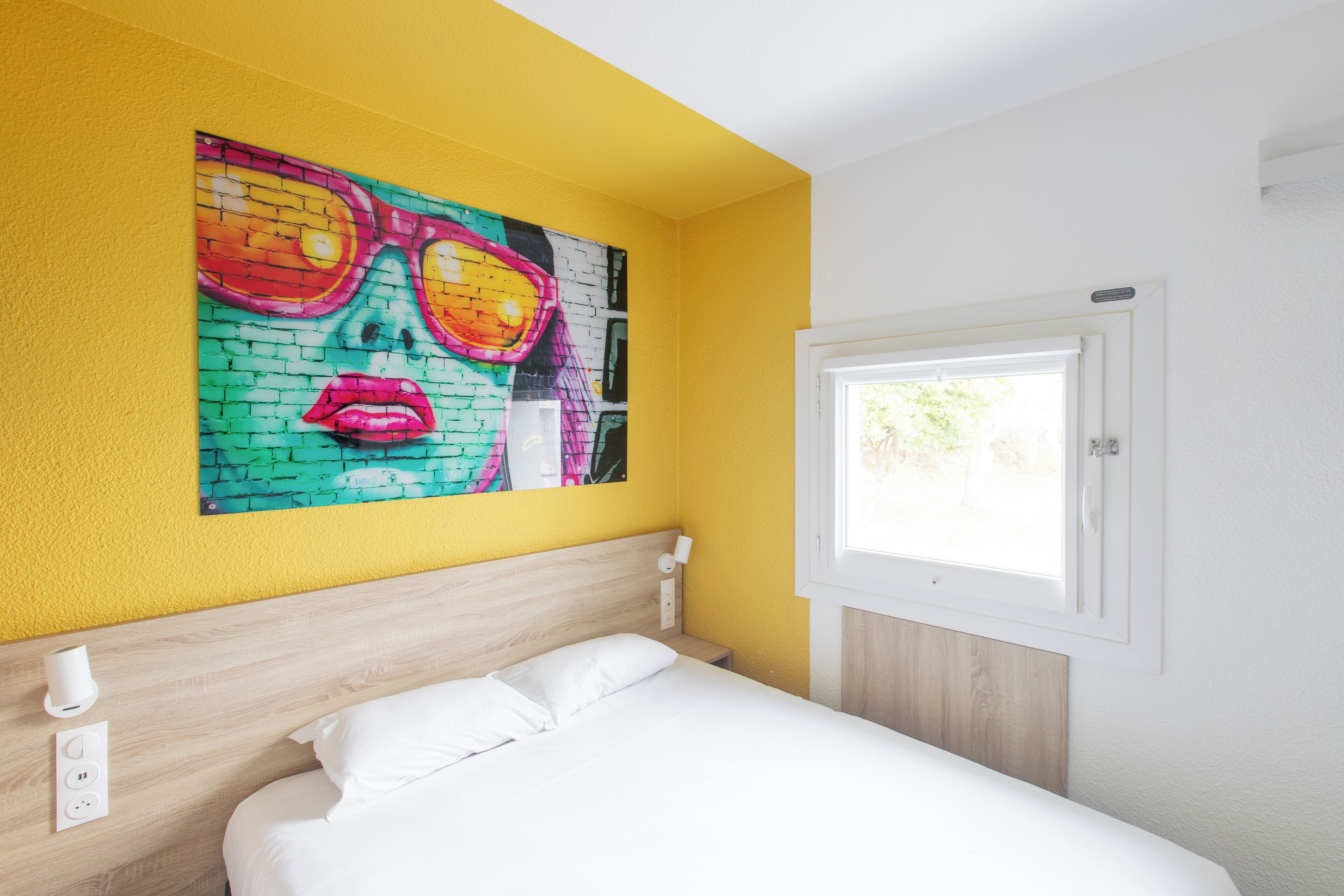 Una habitación de hotel con una cama, un cuadro colgado en la pared, una ventana y una mesita de noche con lámpara.