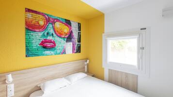 Una habitación de hotel con una cama, un cuadro colgado en la pared, una ventana y una mesita de noche con lámpara.