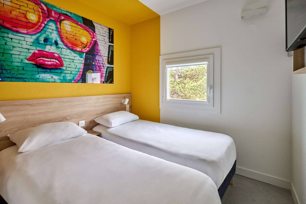 A hotel room with two beds, a colorful mural on the wall, a window with a view of trees, and a TV mounted on the wall.