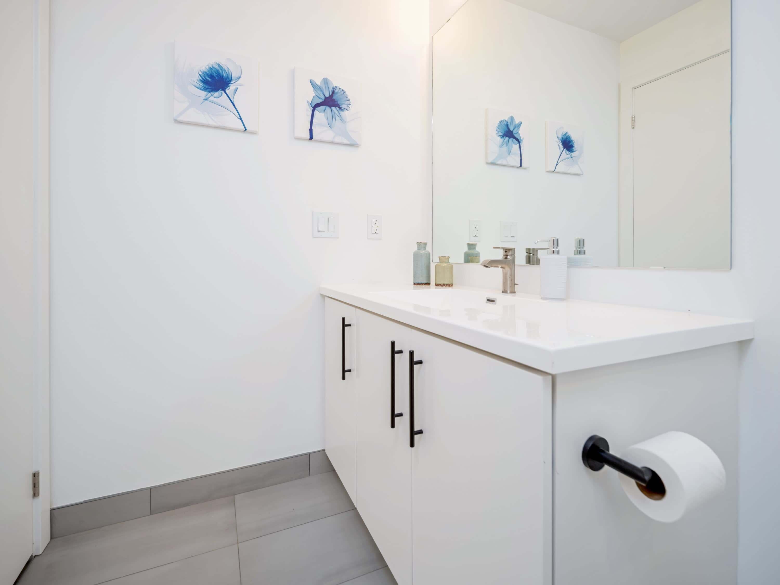 Ein modernes Badezimmer mit einem weißen Waschbecken, Spiegel und Schränken. An der Wand hängen drei gerahmte Blumenbilder.