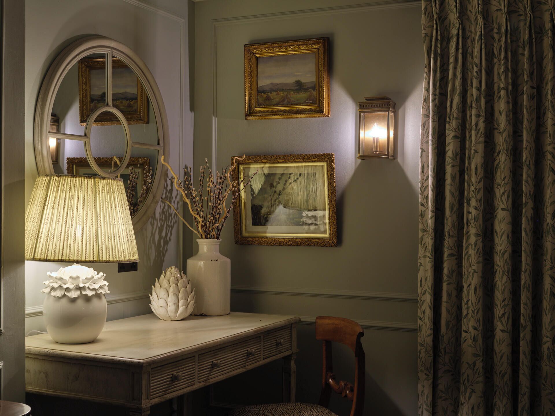 Et værelse med et spejl, en lampe, en vase med grene, et indrammet billede og en vægmonteret lampe.