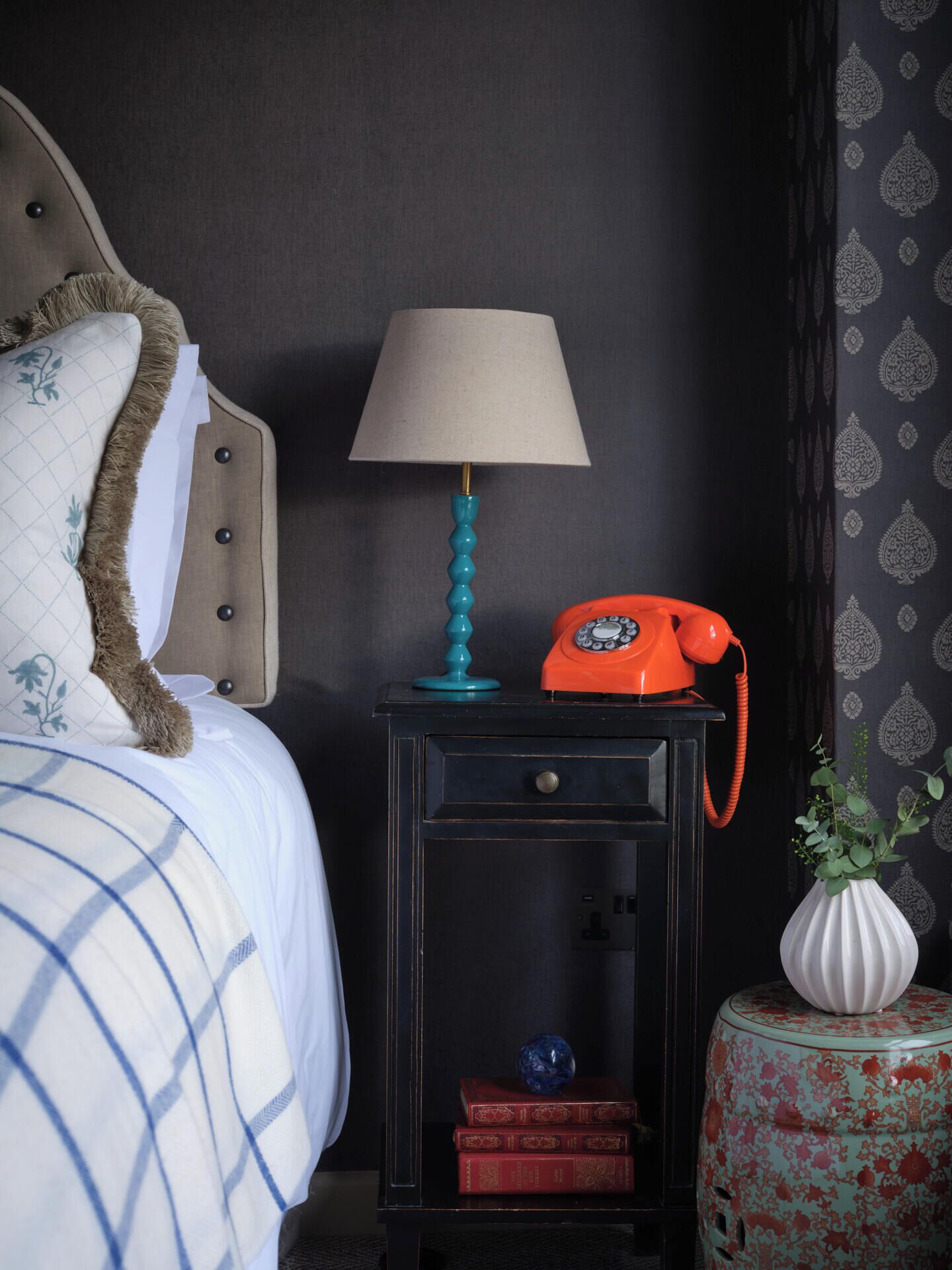 En pænt redt seng med knægt, et lille natbord med lampe, en orange telefon og en dekorativ vase.