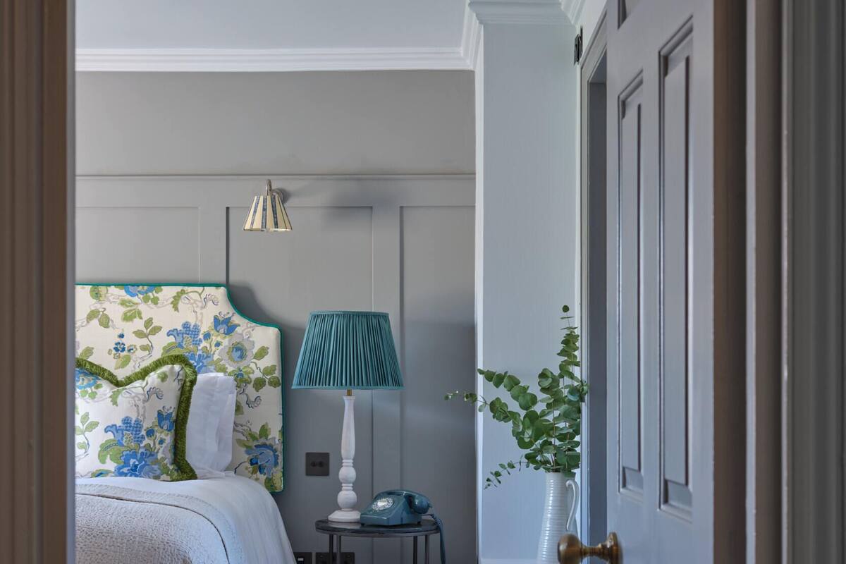 A bedroom with a bed, a bedside table with a lamp, a telephone, and a potted plant.