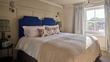 Ein ordentlich bezogenes Bett mit blauem Kopfteil, weißer Bettwäsche und gemusterten Kissen. Zwei Nachttischlampen, ein kleiner Tisch mit einem Radio und ein Fenster mit Vorhängen sind vorhanden.
