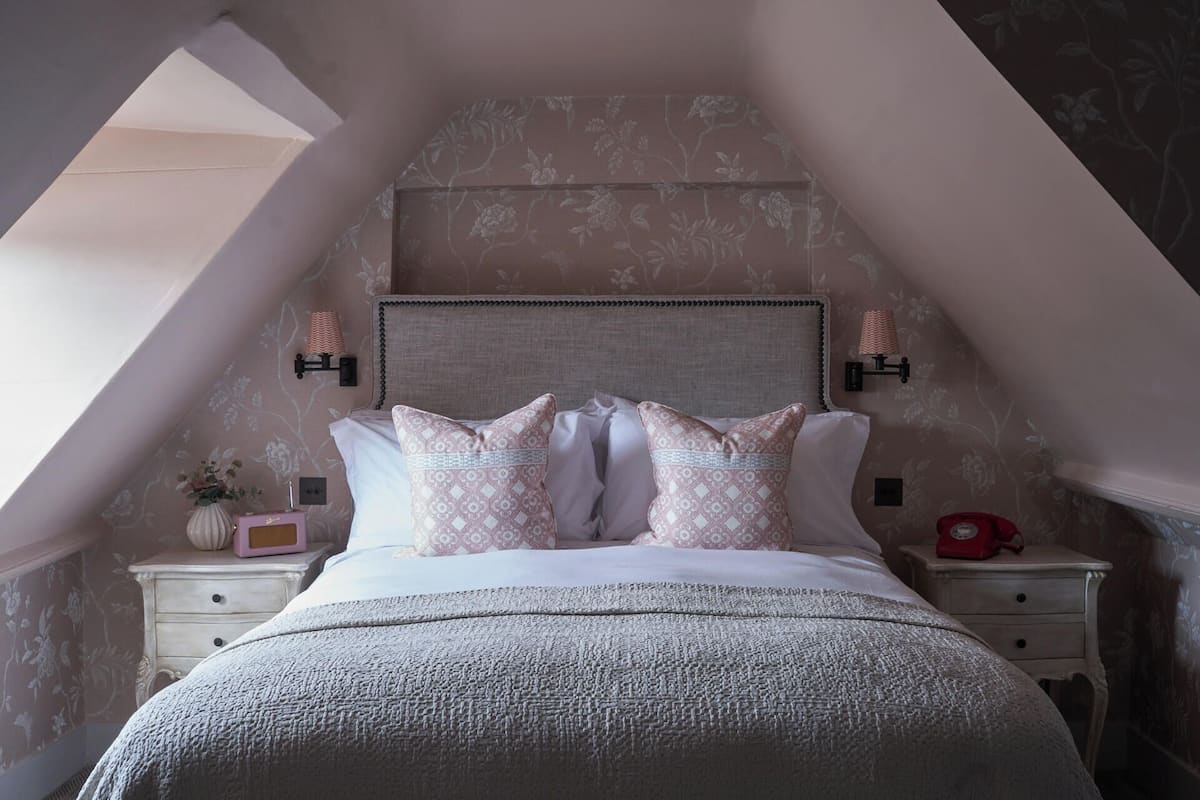 A bedroom with a sloped ceiling, a bed with patterned pillows, two bedside tables with lamps, and a floral wallpaper design.