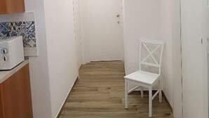 A narrow hallway with wooden flooring, white walls, and a white chair. There is a microwave on a counter to the left.