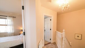 A bedroom with a bed, a door leading to another room, a staircase with a white railing, and a chandelier. - Owen Tucker View House (Ardara)