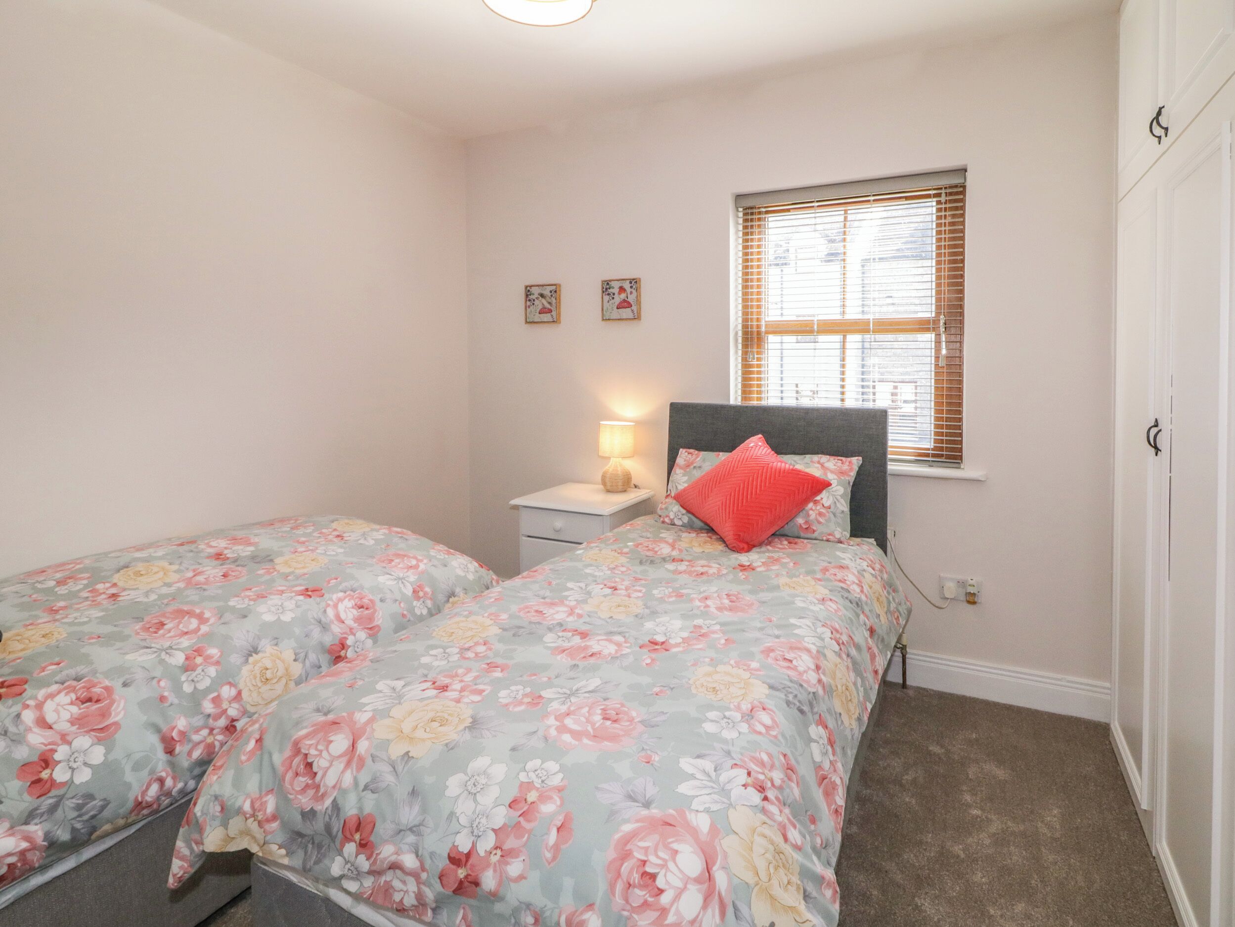 A bedroom with two single beds, a nightstand with a lamp, a window with blinds, and a door.