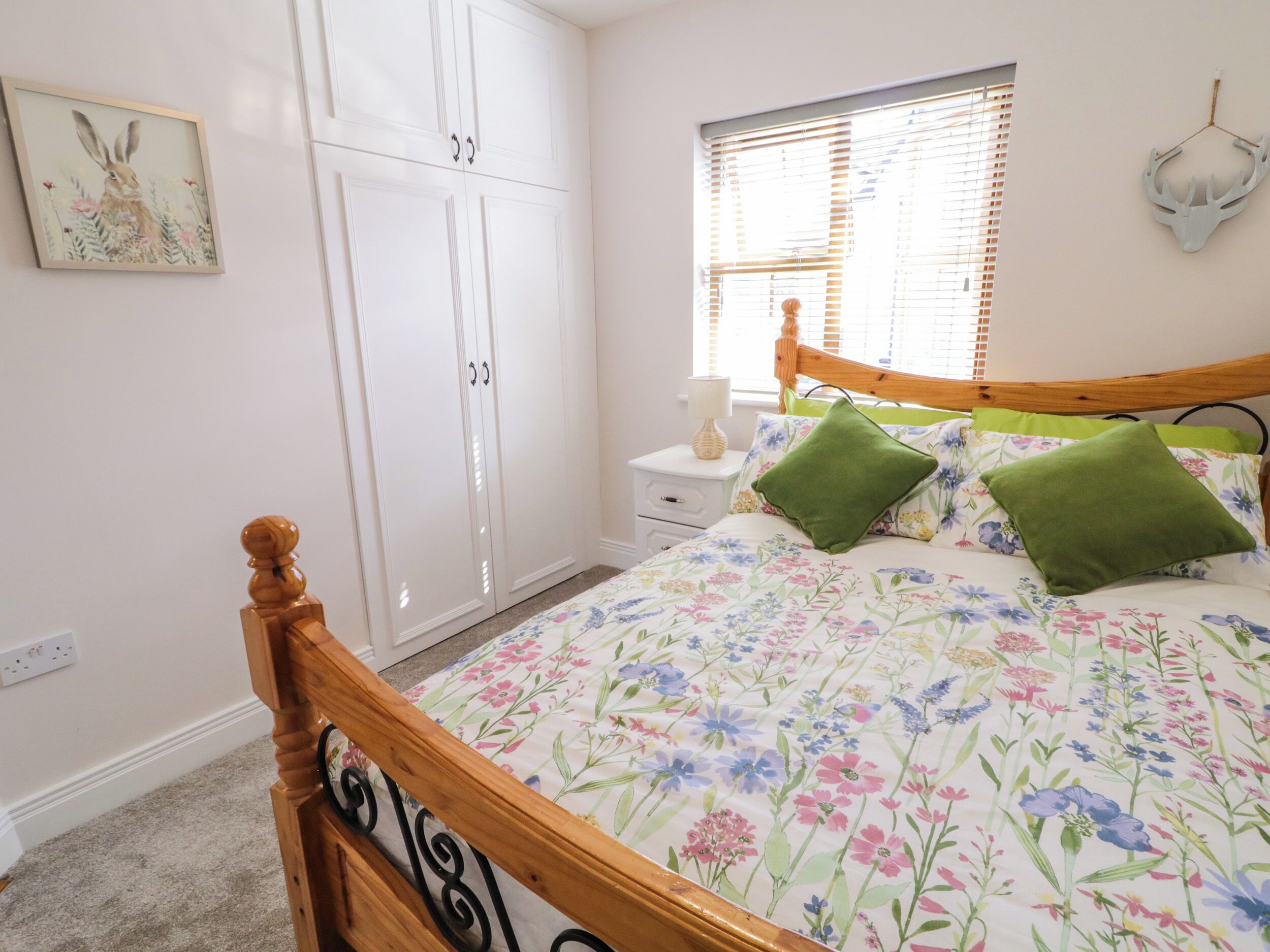 Un dormitorio con estructura de cama de madera, ropa de cama floral, una mesita de noche con lámpara, una ventana con persianas y una decoración de cabeza de venado colgada en la pared.