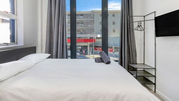 A hotel room with a large bed, a TV mounted on the wall, a window with curtains, and a view of a building outside. - Inn & Out Express Sea Point (Cape Town)