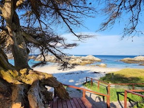 Una terraza de madera que conduce a una playa de arena, un litoral rocoso y un cielo azul despejado.