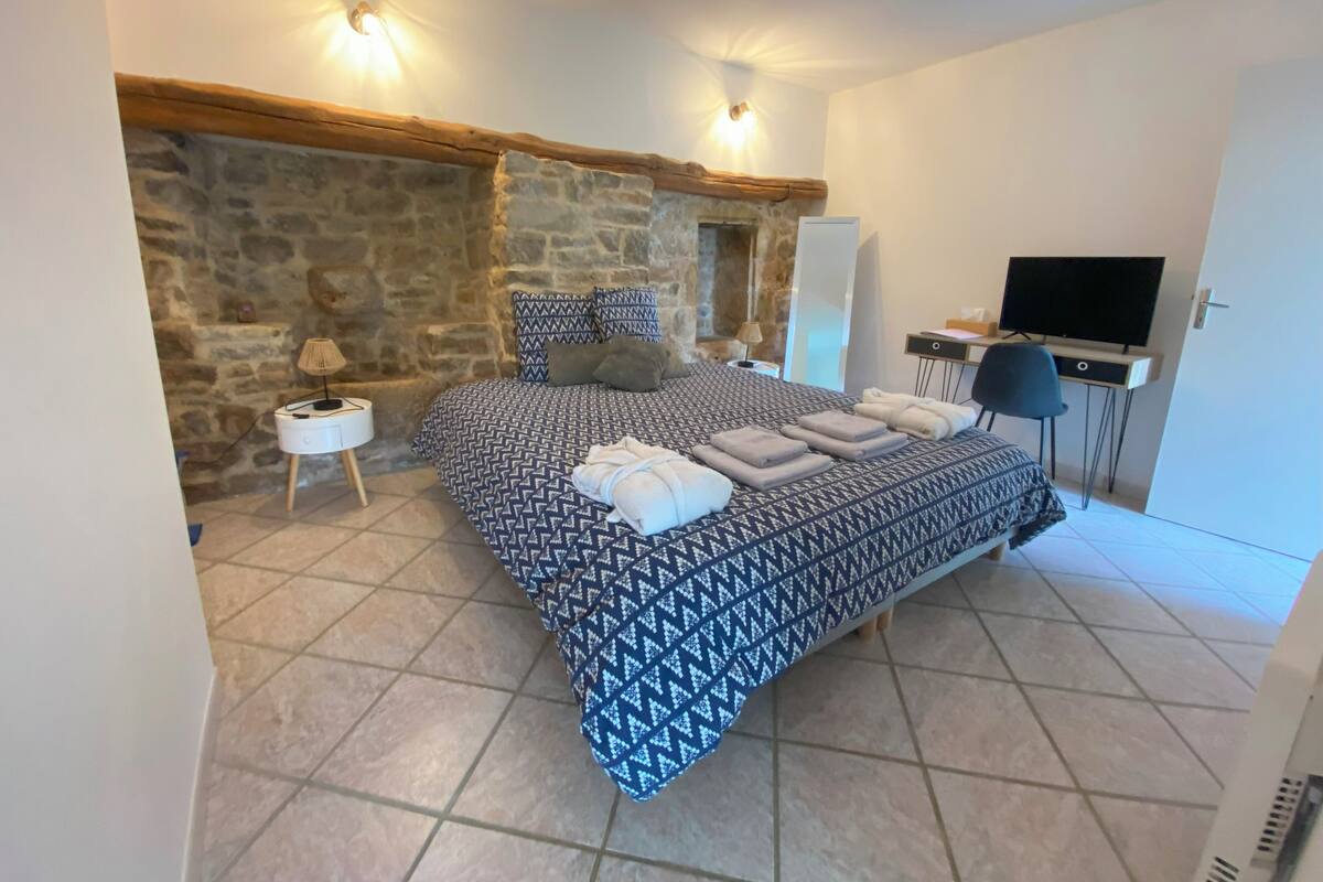 Une chambre avec un mur en pierre, un lit avec une literie à motifs, un bureau avec une télévision et une chaise.