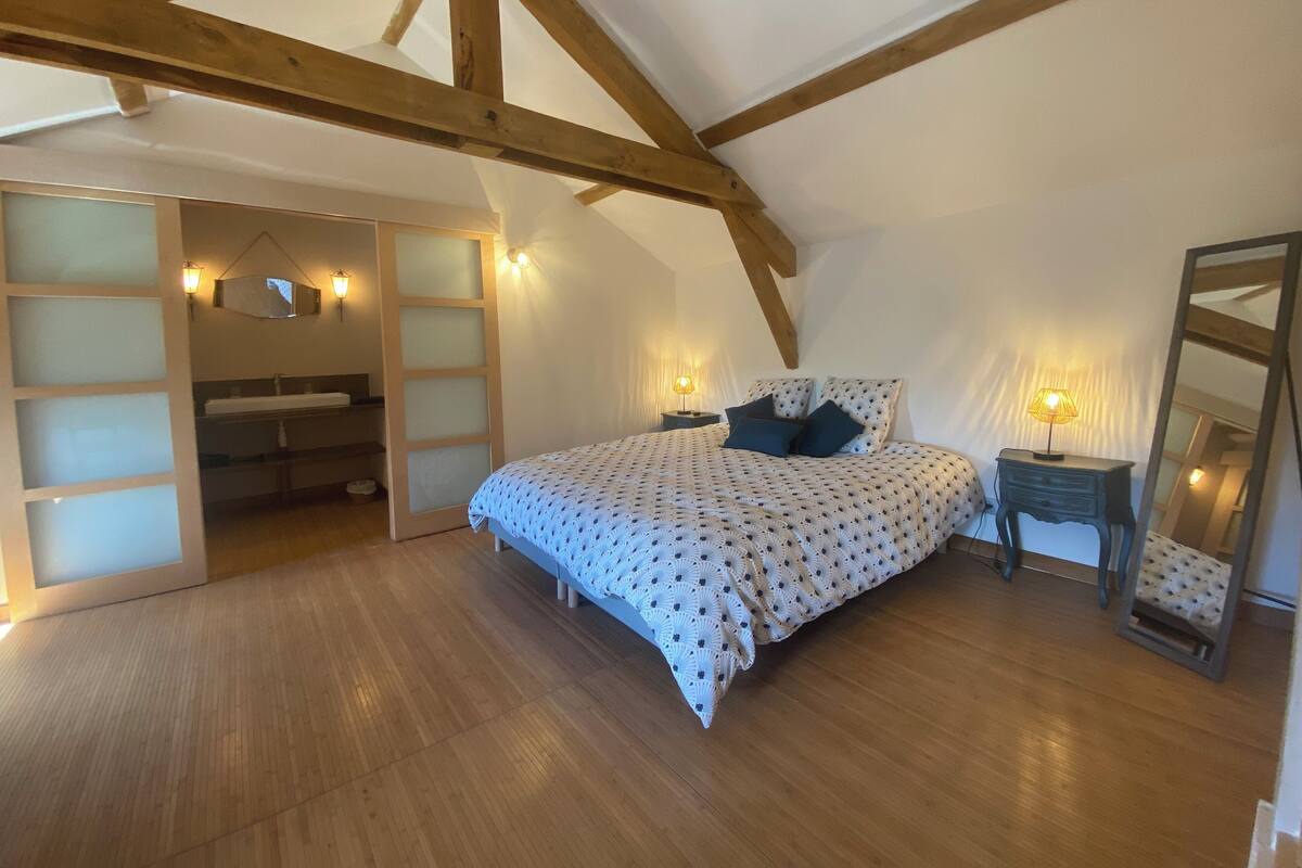 Une chambre avec un plafond en bois, un lit avec une literie à motifs, une table de chevet avec une lampe et un grand miroir.
