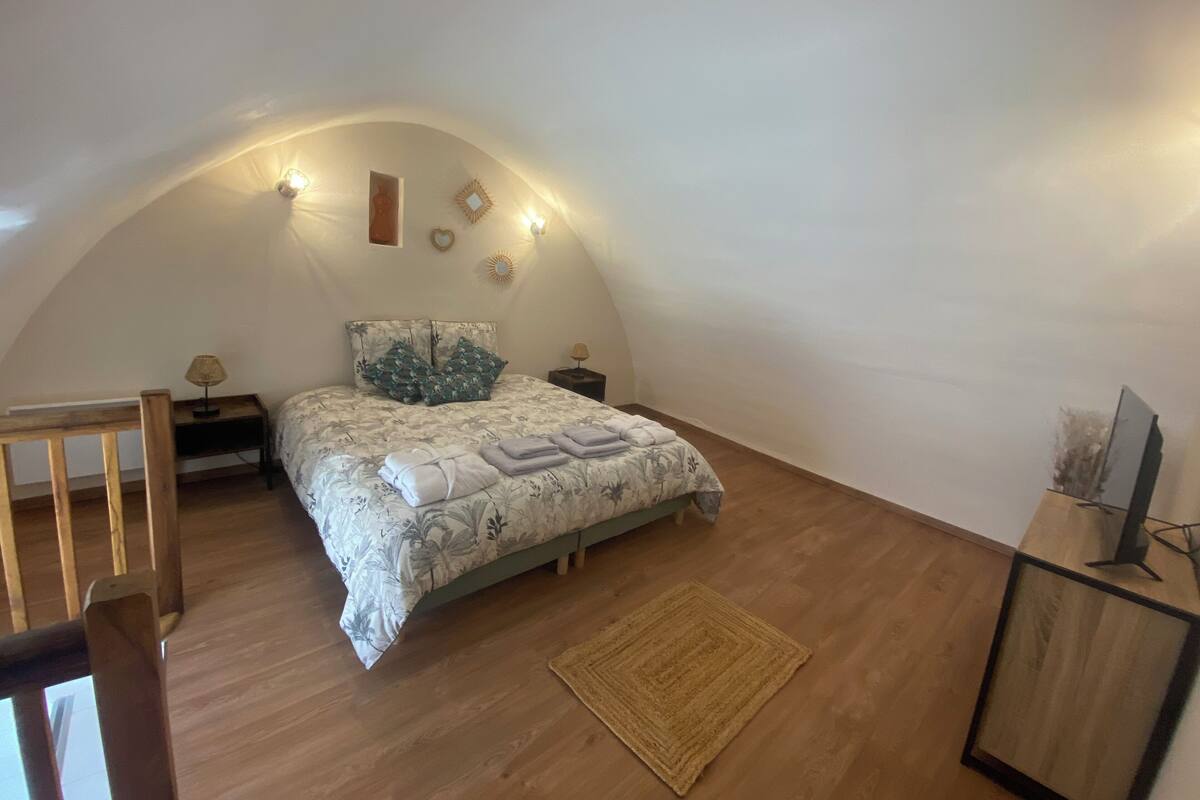 Une chambre avec un plafond en pente, un lit recouvert d’une literie à motifs floraux, une table de chevet avec une lampe et un petit tapis sur le sol.